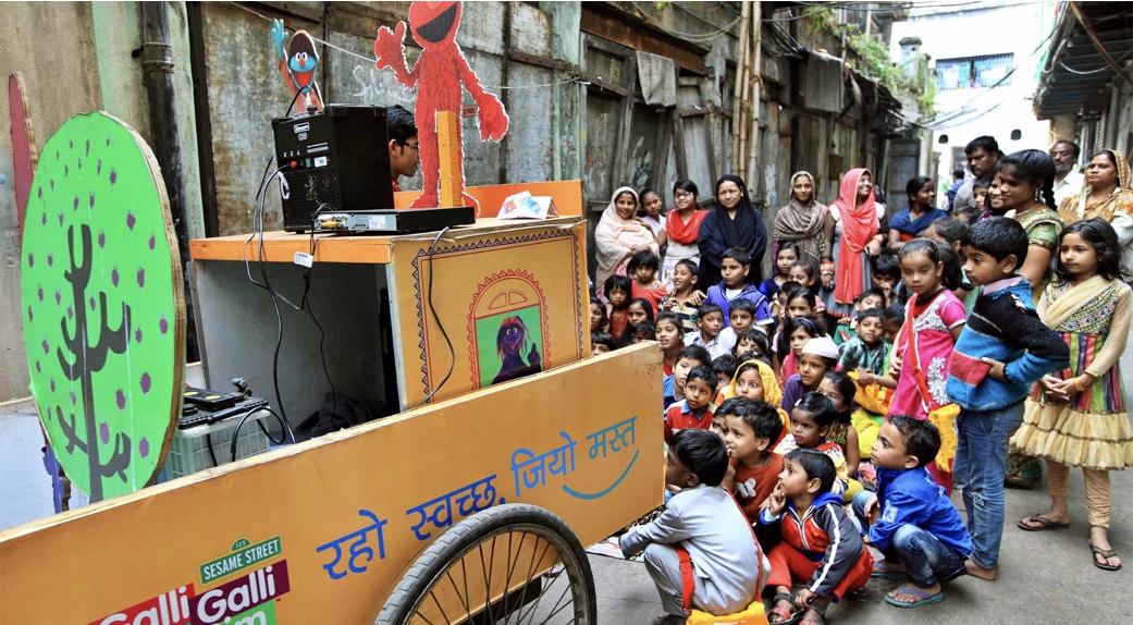 A portable vegetable cart is decorated with sesame street characters, a dvd player, speakers and a tv. Children and their parents gather in an alley way in a urban area. They watch the TV intently.