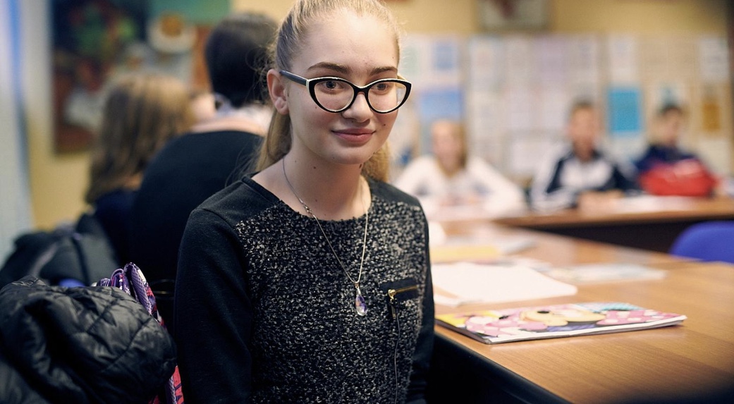 A teenage girl with glasses sits in front of her school desk. There is a publication in front of her.
