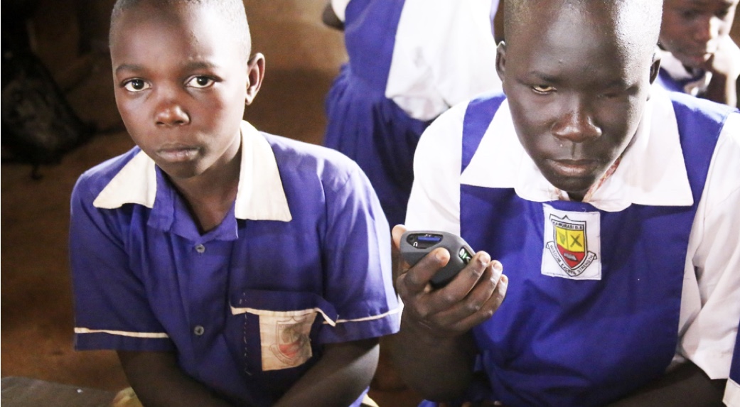 Two children sit in their purple school uniforms. One child has a visual impairment and is using an audio device to learn.