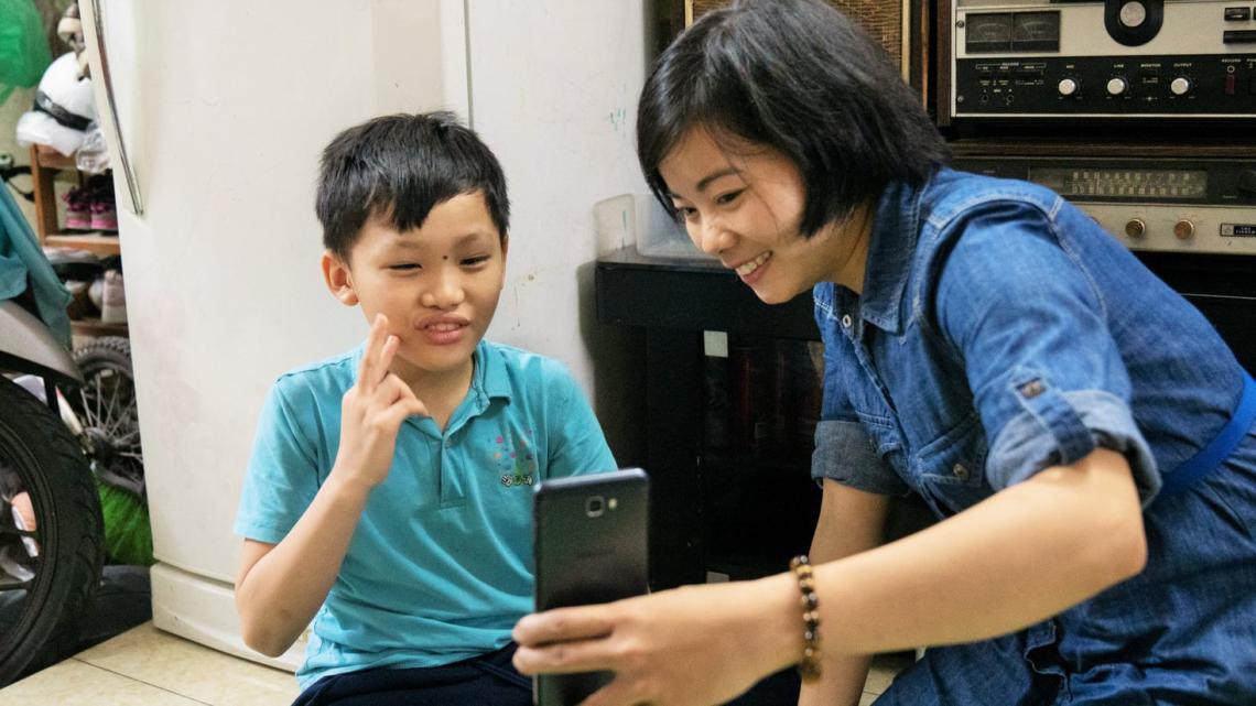 A young boy is signing at the mobile phone. A woman is holding the phone in front of him and smiling.
