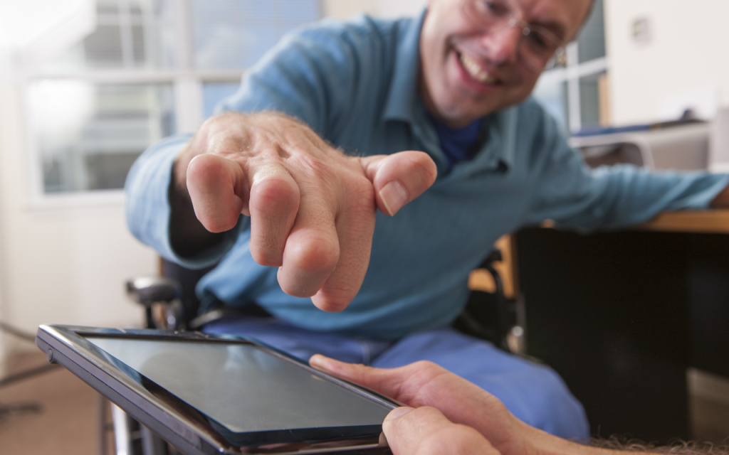 Smiling man in a wheelchair reaches out to touch a tablet, his fingers are curled into a claw.